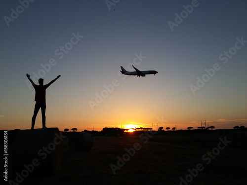 Giovane ragazzo con le mani al cielo e aereo in atterraggio al tramonto