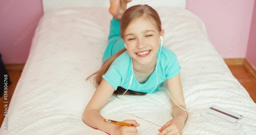 Student doing homework in exercise book. Child lying on bed in bedroom and listening music in headphones and smiling Student doing homework in exercise book. Child lying on bed in bedroom and listening music in headphones and smiling