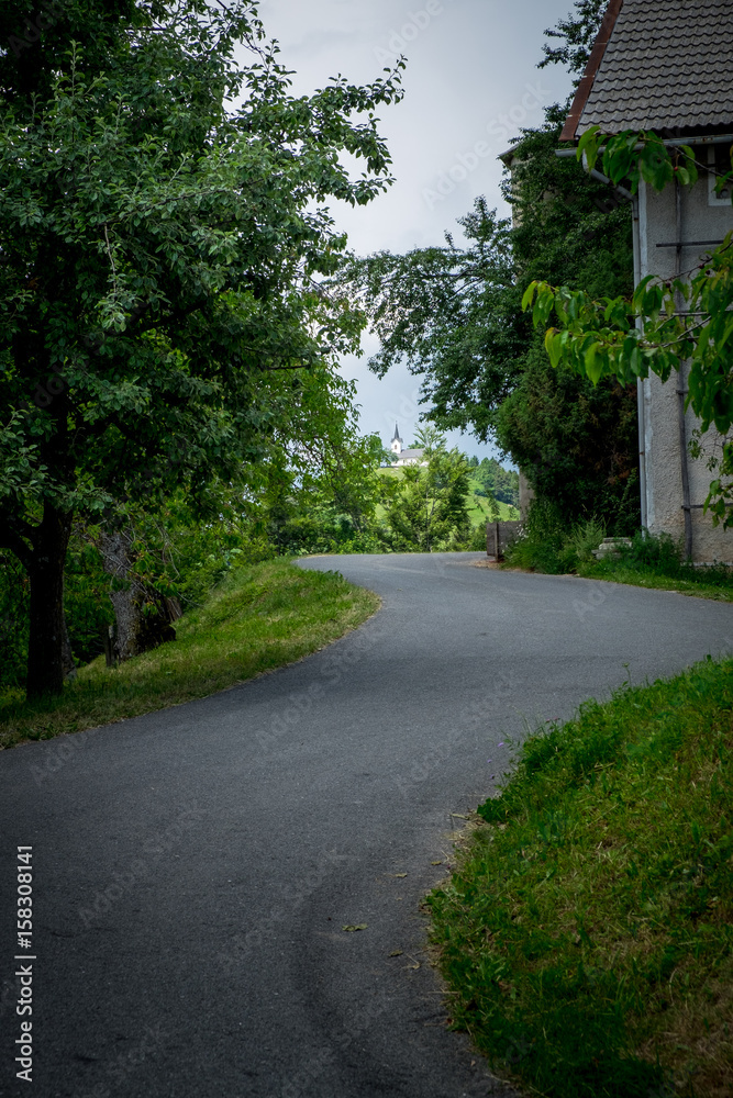 Winding asphalted road leading to the church on the hill. Countryside village landscape with rural house. Rural road inside of village. Winding asphalted road leading to the church on the hill. Countryside village landscape with rural house. Rural road inside of village.