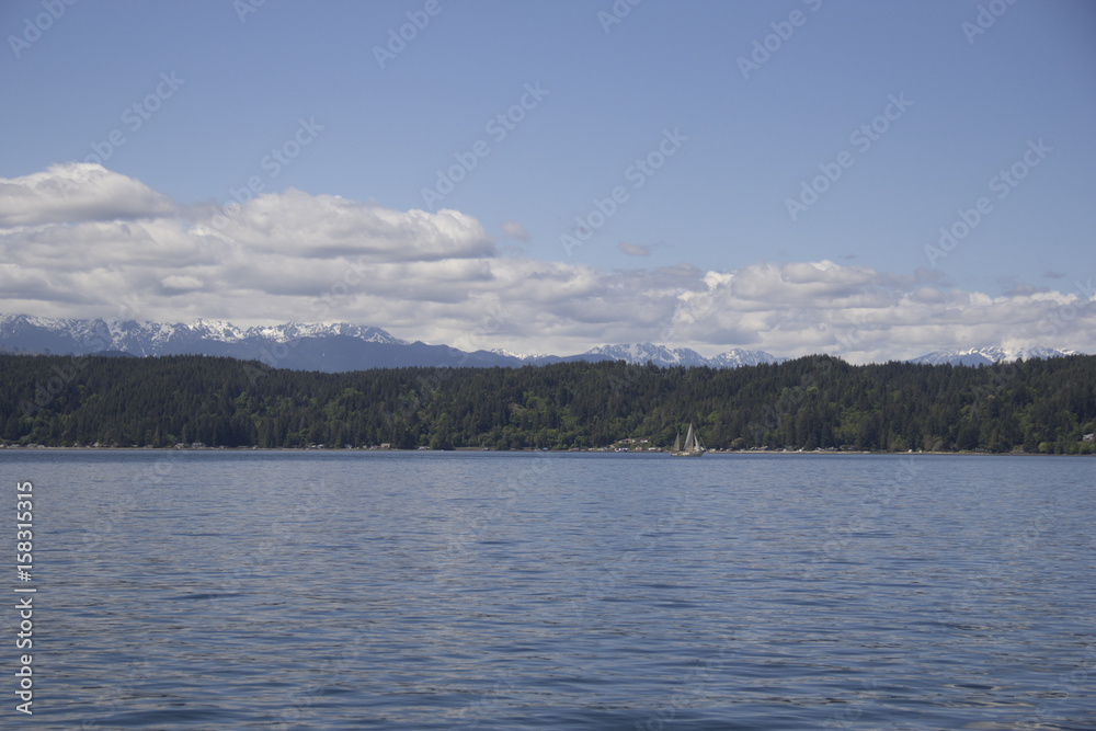 Sailboat sailing in front of forested hills and snow-capped mountains