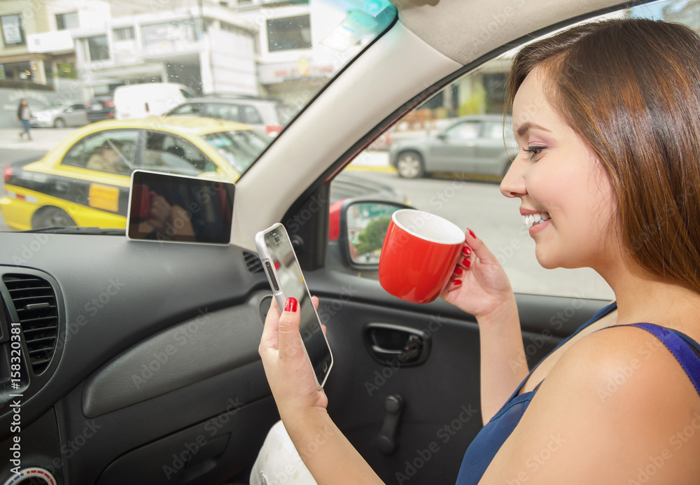 Naklejka premium Close up of a beautiful young woman drinking a cup of coffe or tea and holding in her other hand a cellphone, while the car drives by itself