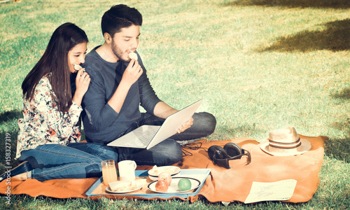 Young happy couple enjoying picnic in park and eating a potato chips while they are watching something in his computer  vintage effect