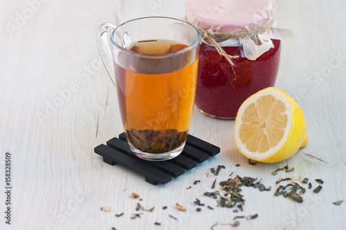 Transparent cup of tea, a jar of raspberry jam, a yellow cut lemon on a wooden stand on a light rustic table. Comfort and warmth. Dry tea around on the table 