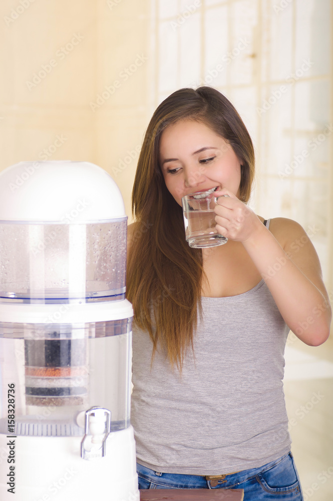 Beautiful woman drinking a glass of water with a filter system of water purifier on a kitchen background Beautiful woman drinking a glass of water with a filter system of water purifier on a kitchen background