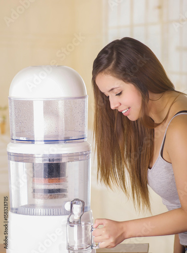 Beautiful smiling woman filling a glass of water, with a filter system of water purifier on a kitchen background