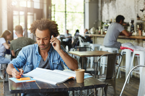 Mindful dark-skinned male student wearing casual clothing preparing for exams sitting at cafe table before lessons, reading information in textbook and talking on phone with his coach asking questions