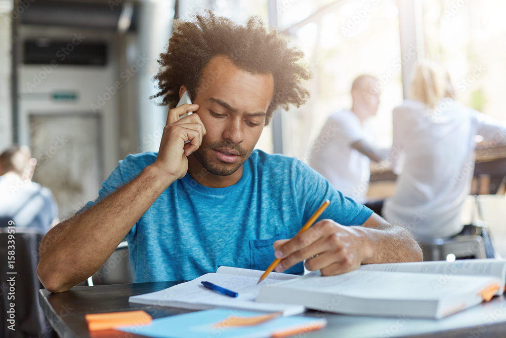 Fototapeta premium Serious African university student having problem doing home task at coworking space, talking on smart phone with groupmate, asking for help, feeling frustrated, surrounded with textbooks and notes