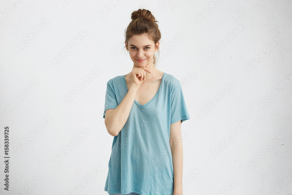 Cute playful young woman with beautiful eyes and charming mysterious smile, touching her chin, having flirting look. Attractive gorgeous girl with hair knot smiling broadly, posing at studio wall Cute playful young woman with beautiful eyes and charming mysterious smile, touching her chin, having flirting look. Attractive gorgeous girl with hair knot smiling broadly, posing at studio wall
