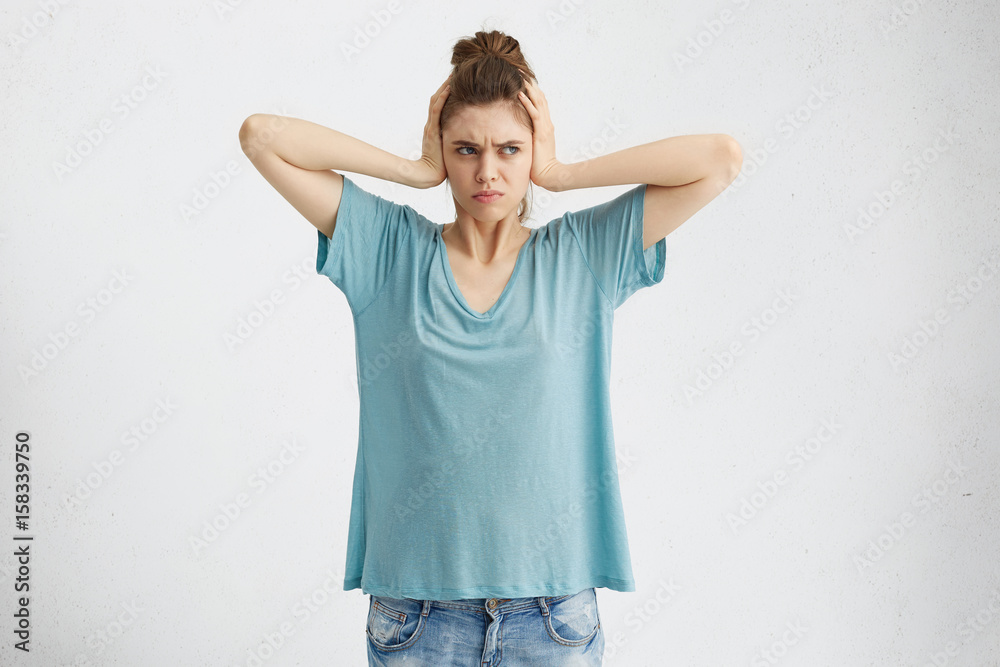 Negative human emotions, reaction and attitude. Frustrated annoyed student girl with hair knot covering ears with hands, feeling irritated with loud annoying noise, can't concentrate on her homework Negative human emotions, reaction and attitude. Frustrated annoyed student girl with hair knot covering ears with hands, feeling irritated with loud annoying noise, can't concentrate on her homework