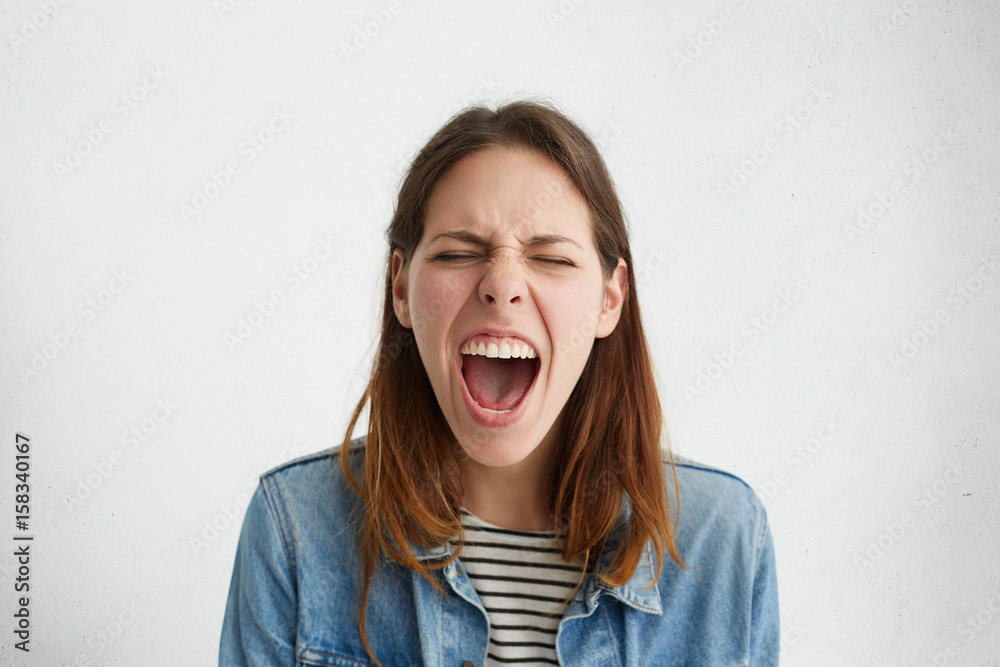 Studio shot of emotional stressed female dressed in trendy clothing shouting in fury and rage, closing eyes tight and opening mouth widely, having mad, furious or fed up look. Human emotions Studio shot of emotional stressed female dressed in trendy clothing shouting in fury and rage, closing eyes tight and opening mouth widely, having mad, furious or fed up look. Human emotions