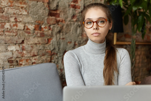 People, modern lifestyle, technology and communication concept. Serious confident young woman entrepreneur in stylish eyewear using laptop pc for remote work, checking e-mail and browsing internet