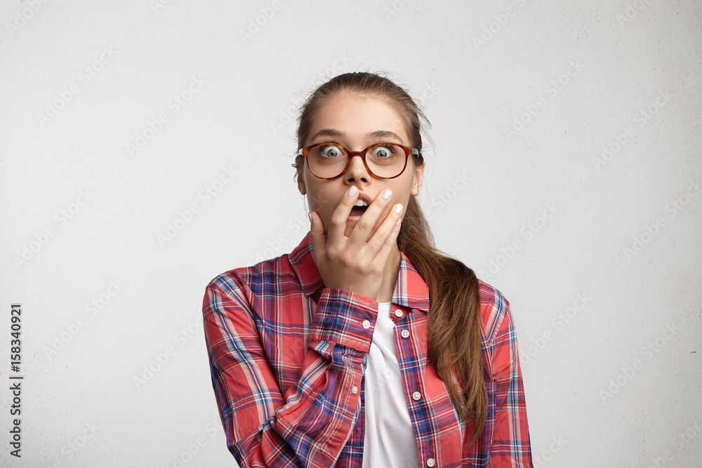 Shocked student female in eyeglasses and elegant shirt looking with wide opened eyes holding hand on mouth being surprised to hear news. Woman in spectacles looking with amazement directly into camera
