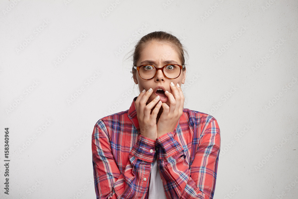 Terrified wide-eyed woman in glass spectacles and red checked shirt holding her hand on opened mouth being scared after what she done. Astonished woman regreting her actions being afraid of mistakes Terrified wide-eyed woman in glass spectacles and red checked shirt holding her hand on opened mouth being scared after what she done. Astonished woman regreting her actions being afraid of mistakes