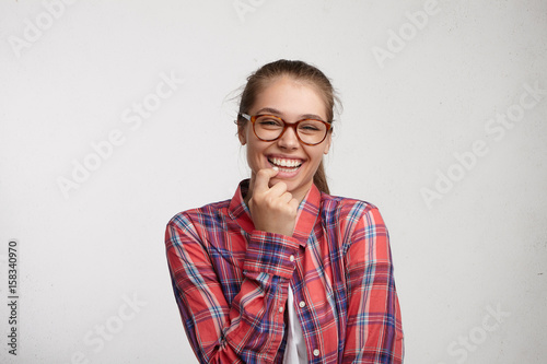 Portrait of attractive pleasant smiling woman with fair hair wearing elegant eyeglasses and checked red shirt holding finger on teeth isolated over white background having good mood after party. © Wayhome Studio