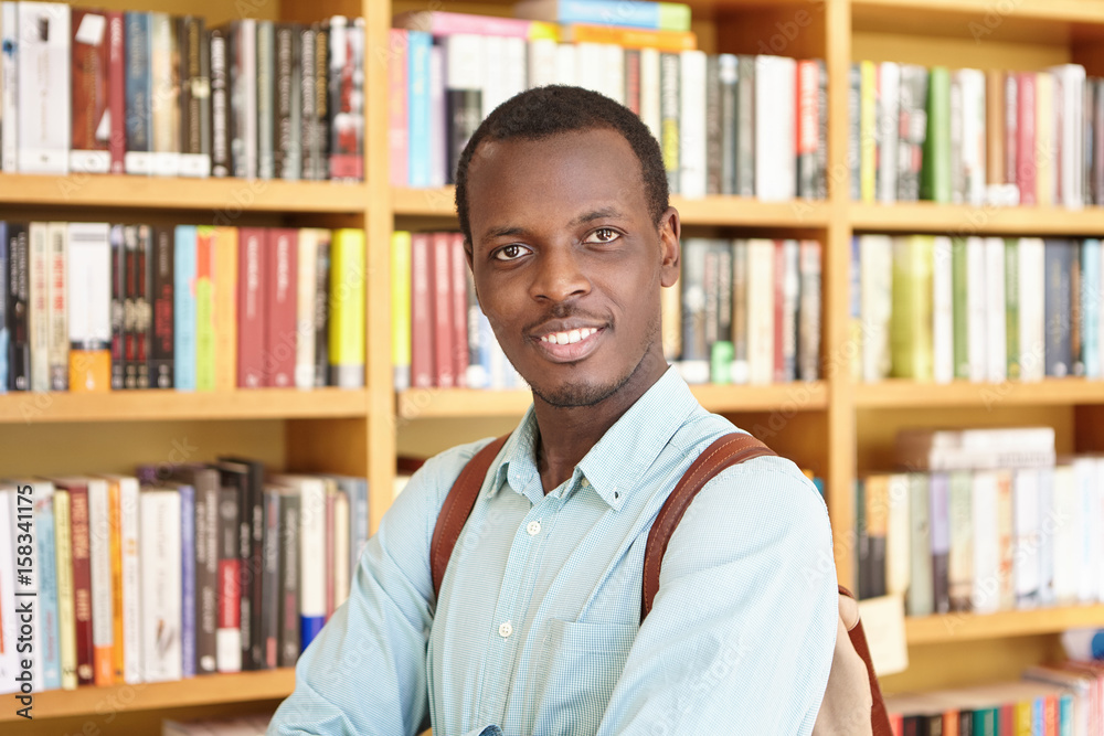 Black college student wearing elegant shirt holding rucksack isolated over library background looking with smile into camera. Male student visiting university library finding necessary book for report Black college student wearing elegant shirt holding rucksack isolated over library background looking with smile into camera. Male student visiting university library finding necessary book for report