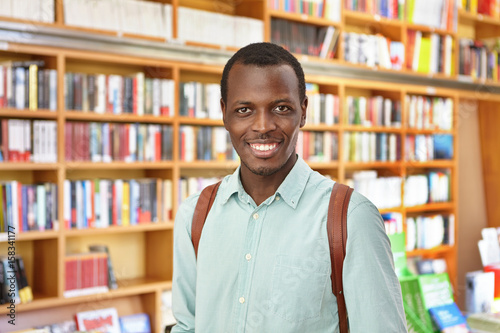 Portrait of dark-skinned male student holding rucksack standing in library choosing interesting book to read having pleasant smile enjoying reading. African student standing against bookshelves