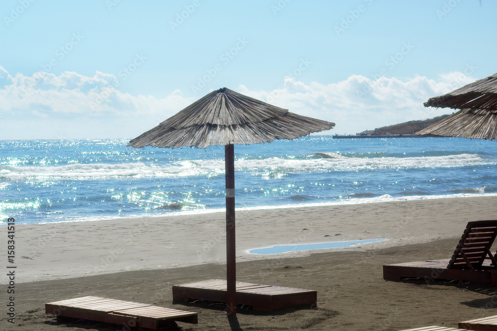 An umbrella from the sun made of reeds and wooden sunbeds stand empty on the beach, sparkling sea with waves