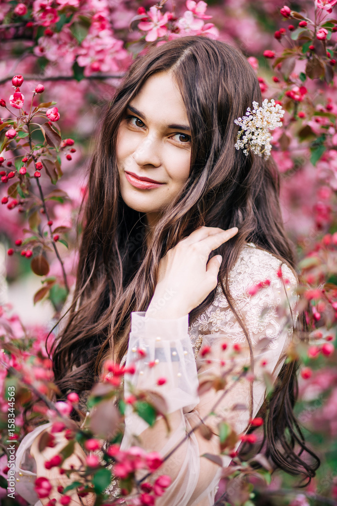 beautiful cute brunette girl bride in boudoir lace transparent dress, with decoration on the hair, smiling, looking around the tree blossoms with pink flowers