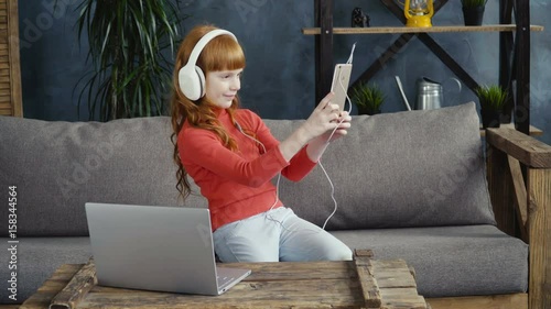 Little girl in headphones is making a selfie on the sofa in the room