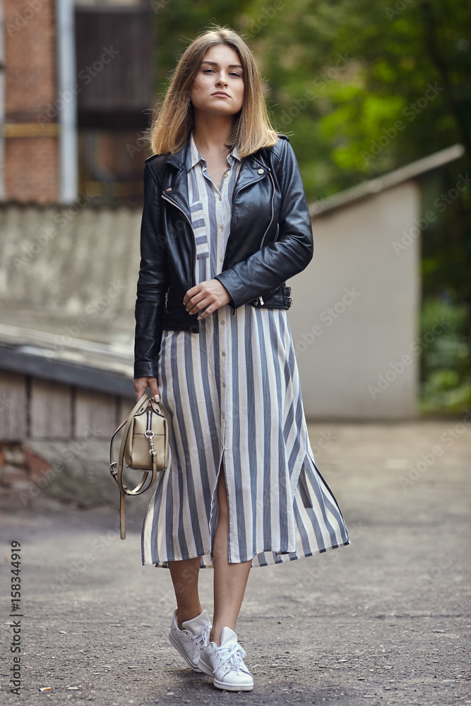Young beautiful girl in stylish streetwear black leather jacket long striped dress white sneakers and with a fashionable bag strolling in summer city park