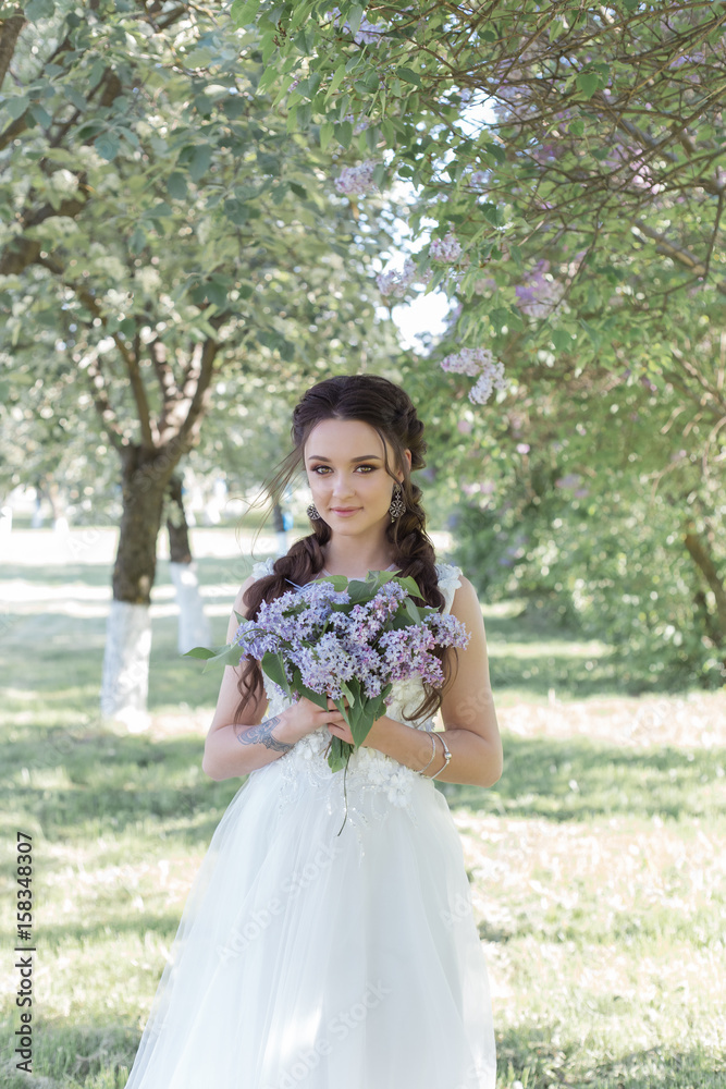 Tender beautiful cute girl bride in a white air dress with a bouquet of lilacs in her hands walking through the park on a sunny spring day. Photo in gentle colors