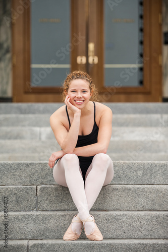 Beautiful and young ballerina smiling while seated on concrete steps at bottom of frame.  