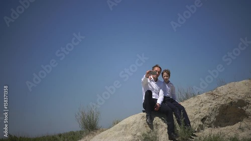 Happy father and son are photographed against the sky, sit on a hill, selfie