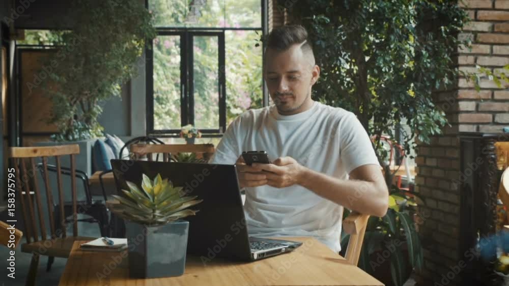 Caucasian young businessman answer messages on smartphone. Man work on laptop and mobile phone in cosy cafe Caucasian young businessman answer messages on smartphone. Man work on laptop and mobile phone in cosy cafe