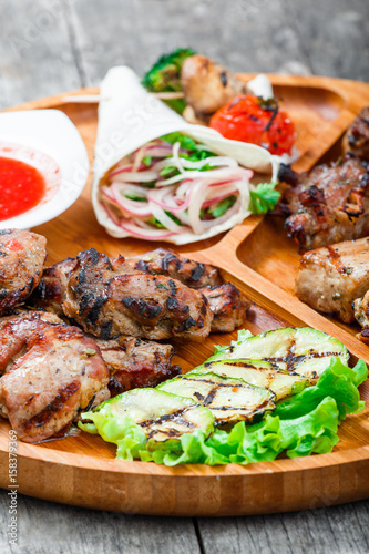 Assorted delicious grilled meat and vegetables with fresh salad and bbq sauce on cutting board on wooden background close up