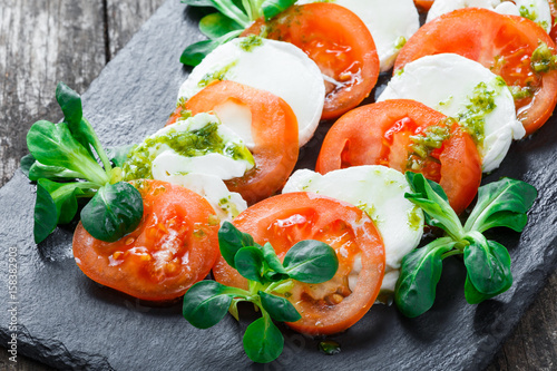 Caprese salad with Mozzarella cheese, tomatoes, pesto sauce and basil herb leaves on black slate stone chalkboard on background
