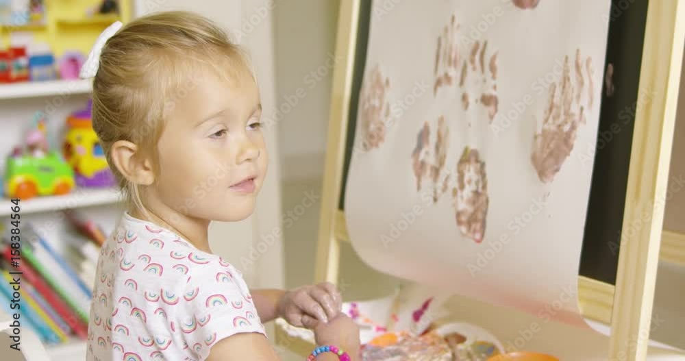Happy attractive little blond girl artist rubbing her ands with a cute smile after applying palm prints to her abstract painting on an easel black board at home or in kindergarten. Happy attractive little blond girl artist rubbing her ands with a cute smile after applying palm prints to her abstract painting on an easel black board at home or in kindergarten.