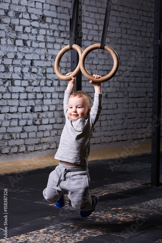 Cute child boy hanging on gymnastic rings in the cross fit gym against brick wall.