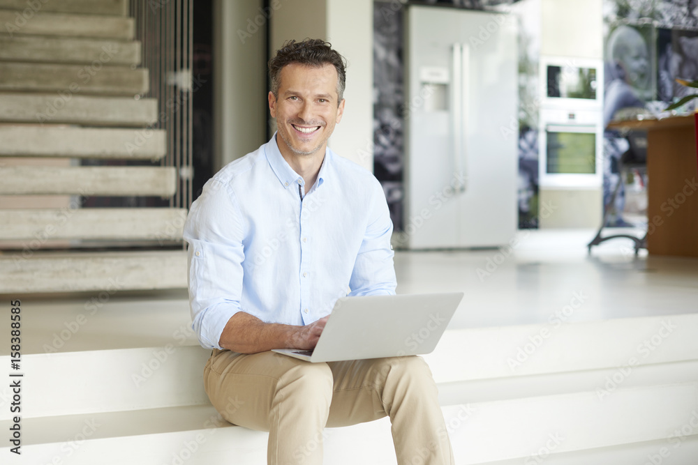 Manage my business from home. Shot of a smiling casual man using his laptop and working online while sitting on stairs at his modern home. Manage my business from home. Shot of a smiling casual man using his laptop and working online while sitting on stairs at his modern home.