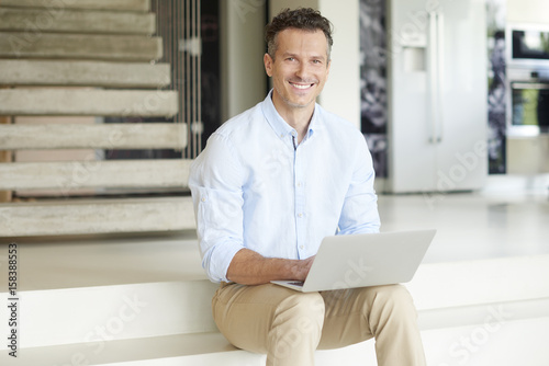 Manage my business from home. Shot of a smiling casual man using his laptop and working online while sitting on stairs at his modern home. 