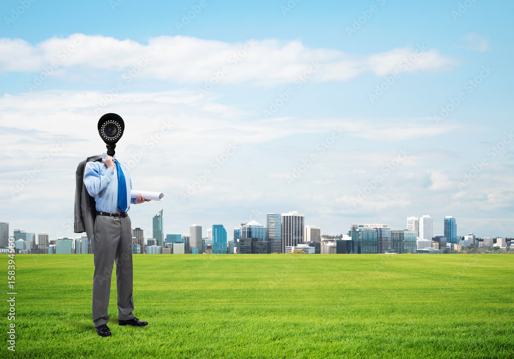 Camera headed man standing on green grass against modern cityscape Camera headed man standing on green grass against modern cityscape
