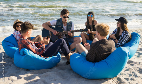 group of friends with guitar on the beach party - Young hipster people on summer vacation sitting on bean bags and playing guitar near the sea