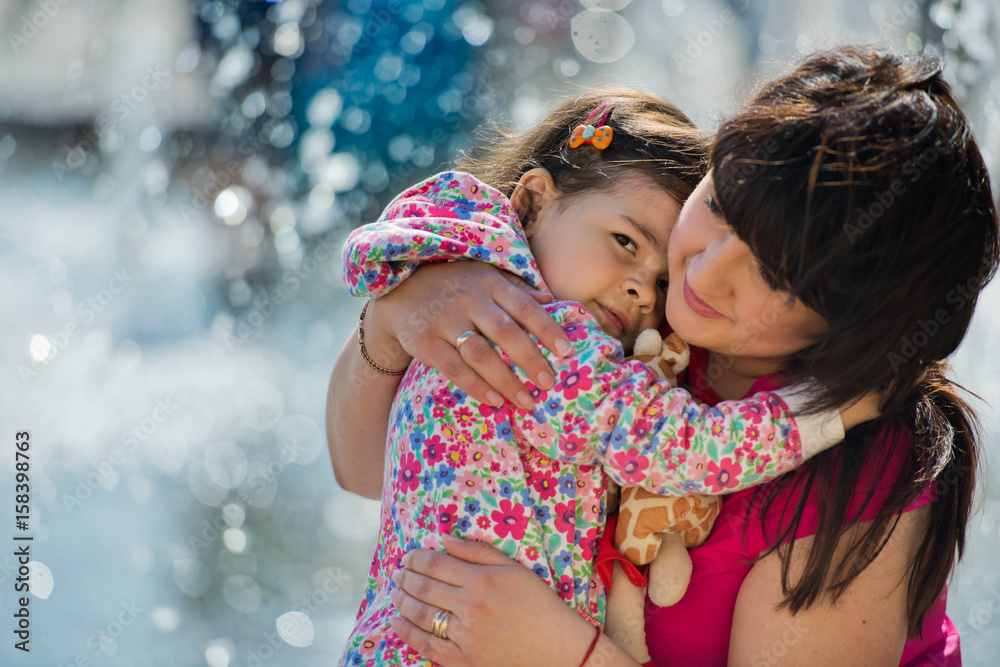 Obraz premium A small charming girl hugs her mother in the park near the fountain. Mothers Day.