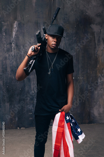 Armed confident black man with american flag and weapon stay on dark background. Gangster, patriotism, social problem, immigration, us citizenship, armed strike, bandit groups concept