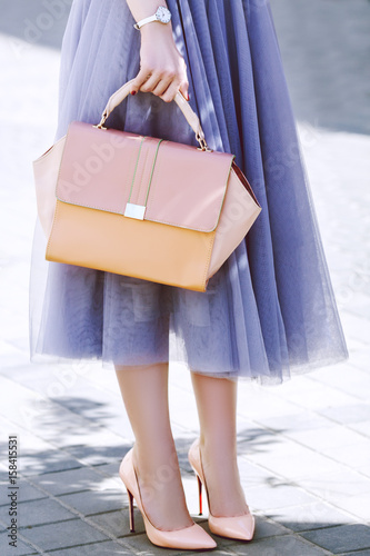 Fashionable woman posing in street. Girl holding elegant pink bag, purse, wearing stylish lash tulle skirt, shoes, wrist watch. Luxury wear and accessories. City lifestyle. Female fashion concept
