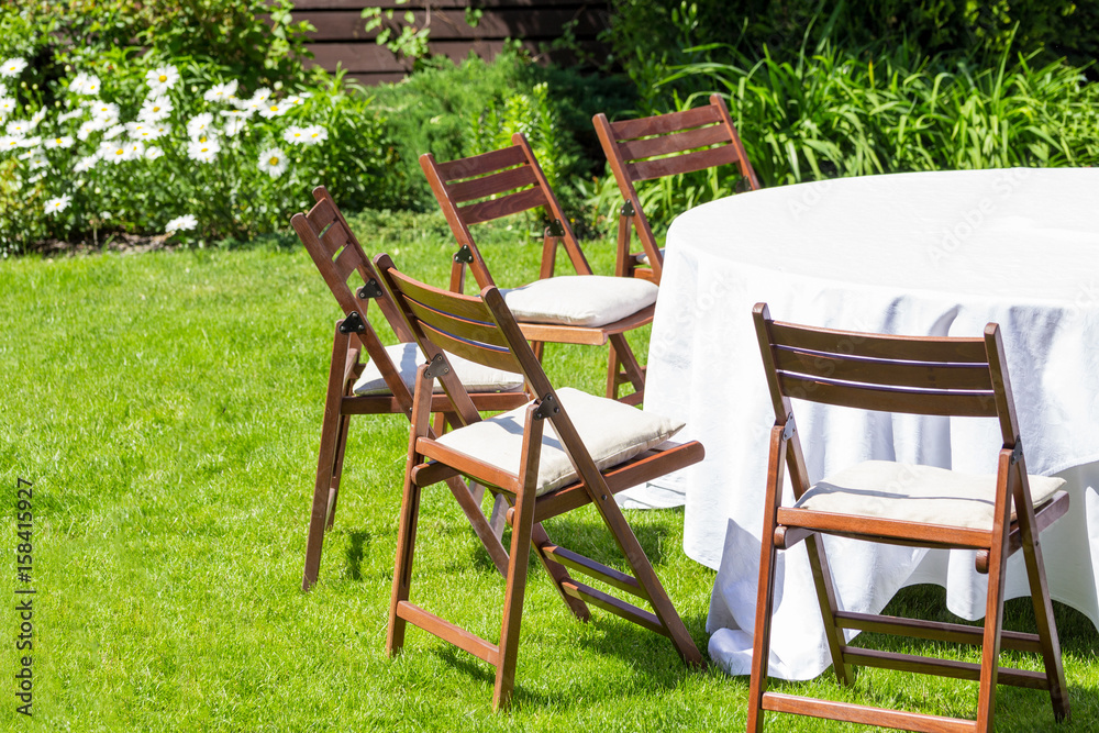 Round table covered with white cloth and chairs stand on a green lawn outdoors .