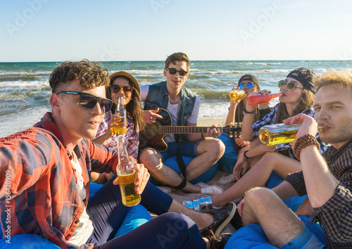 cheerful group of friends taking selfie and having fun at the beach. They play guitar and drink alcohol. Happy youth time. Sea is on background © Igor Kardasov
