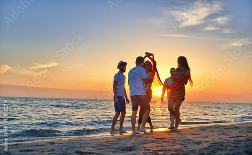 group of happy young people dancing at the beach on beautiful summer sunset