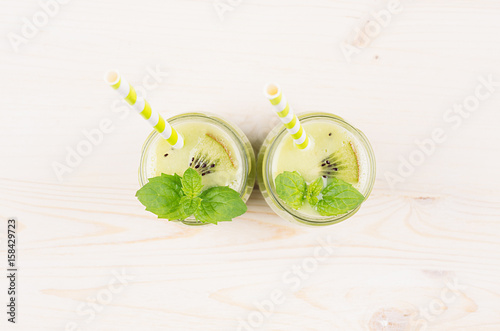 Freshly blended green kiwi fruit smoothie in glass jars with straw, mint leaf, top view. White wooden board background, copy space.