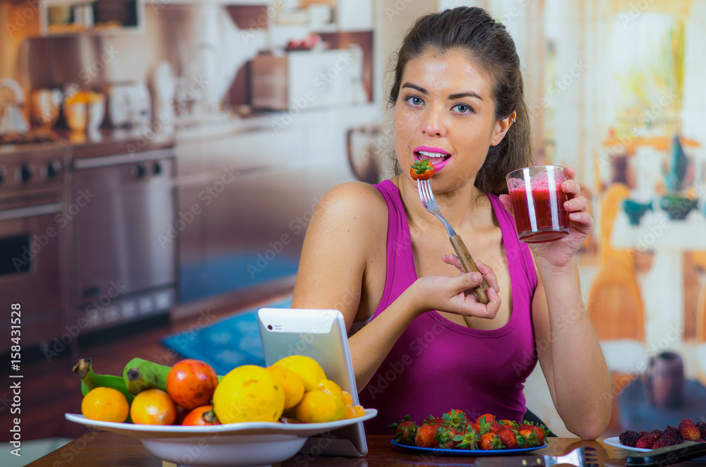 Young woman wearing pink top enjoying healthy breakfast, eating strawberries and smiling, home kitchen background