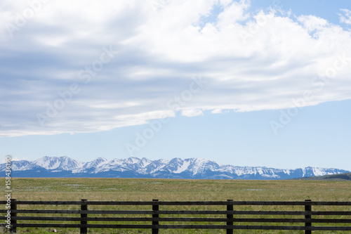 Wooden fence and prairie in the foreground with rugged snow capped mountains in the background. Fluffy gray and white clouds against light blue sky are above.