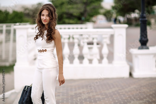 Beautiful stylish brunette girl, street portrait, street fashionable look. Beautiful woman with long hair in white clothes and a handbag in her hands, a strict style, walking on the street