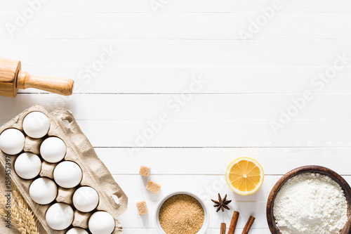 Baking ingredients on white table. Box of white eggs, brown sugar, spices, lemon, white flour and rolling pin on white wooden table background. Top view and copy space