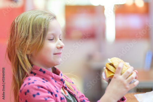 Adorable blonde girl in pink clothes eat double burger with beef and cheese in fast food restaurant
