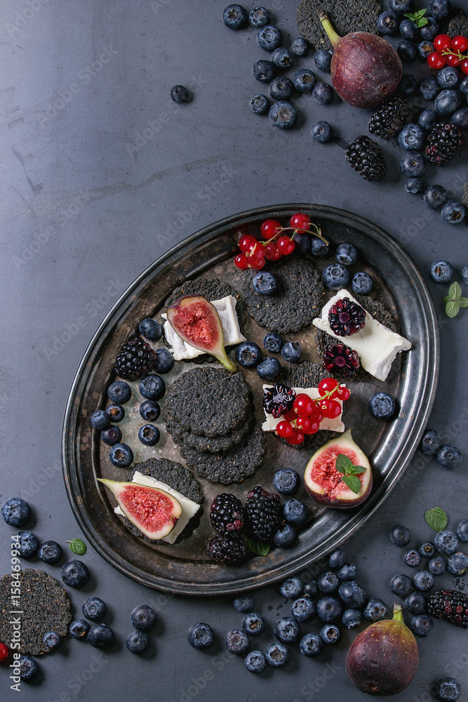 Black charcoal crackers with camembert brie cheese and berries blueberry, dewberry, red currant and sliced figs, served on vintage tray over dark metal background. Top view, space. Summer appetizer Black charcoal crackers with camembert brie cheese and berries blueberry, dewberry, red currant and sliced figs, served on vintage tray over dark metal background. Top view, space. Summer appetizer