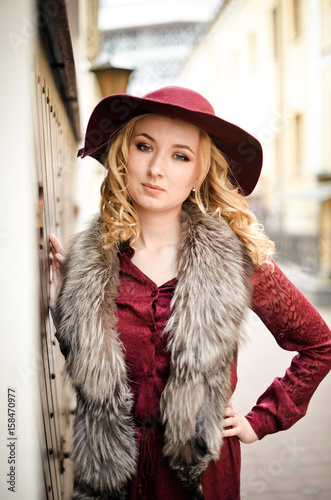 Красивая девушка со светлыми волосами стоит на улице в бордовом платье и шляпе с меховым воротником 